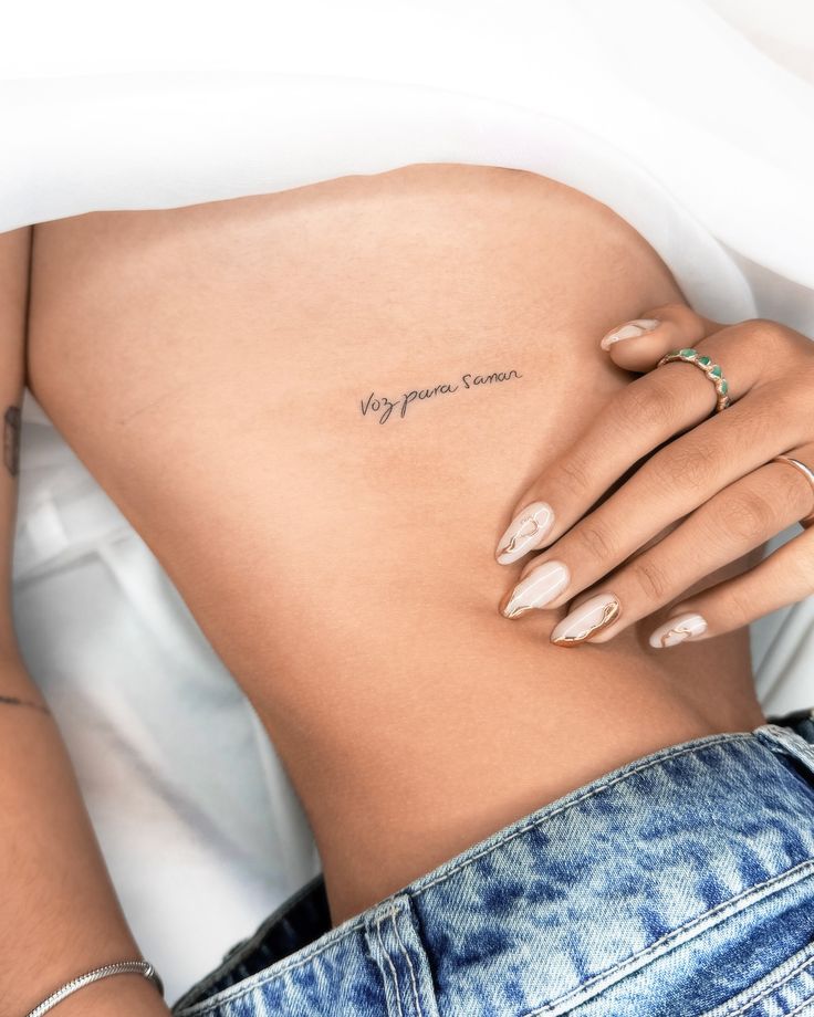 Close-up of a minimalistic script tattoo on a person's side, with manicured nails and blue jeans visible.