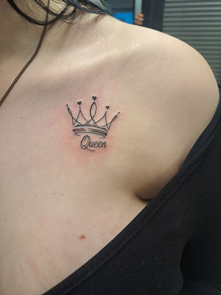 tattoo Elegant crown tattoo with Queen text on woman's shoulder. Minimalist design on skin, emphasizing style and empowerment. | Sky Rye Design Elegant crown tattoo with Queen text on woman's shoulder. Minimalist design on skin, emphasizing style and empowerment.