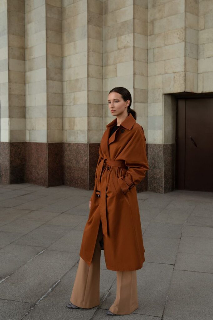 Elegant woman in brown trench coat, standing by stone building exterior.