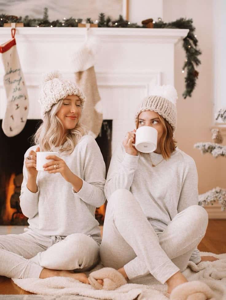 Two women in cozy pajamas enjoying hot drinks by a decorated fireplace during the holiday season.