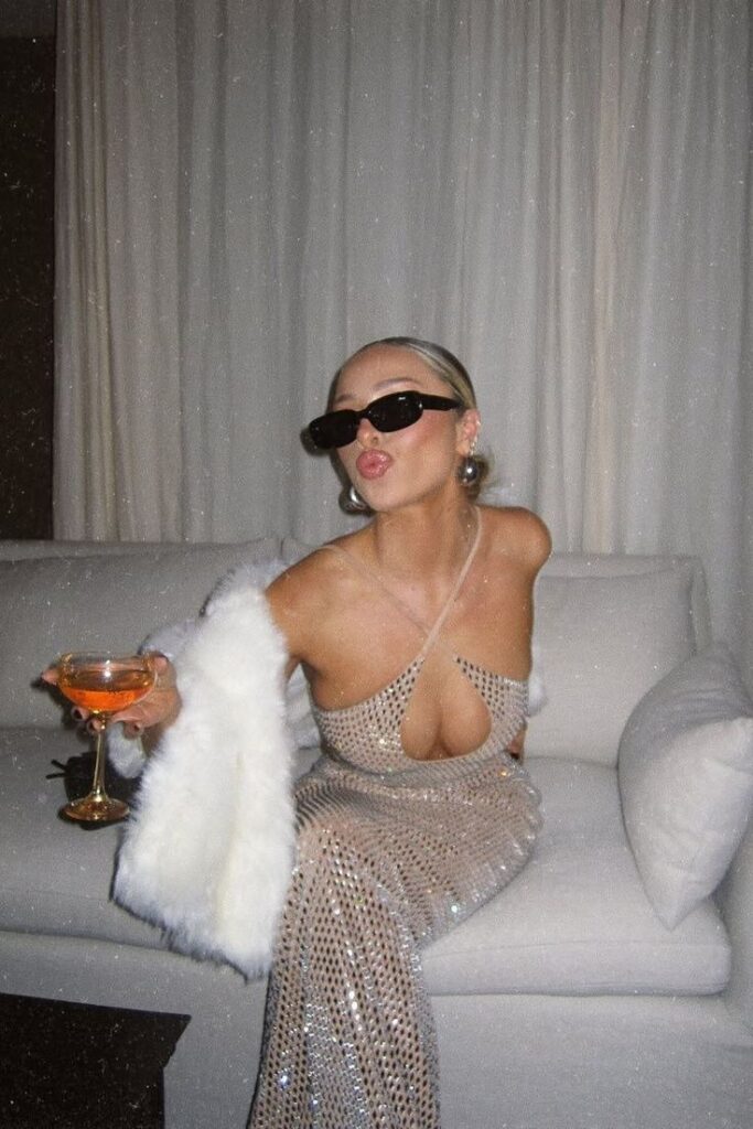 Woman in sparkling gown and sunglasses, holding a cocktail and posing glamorously on a couch.