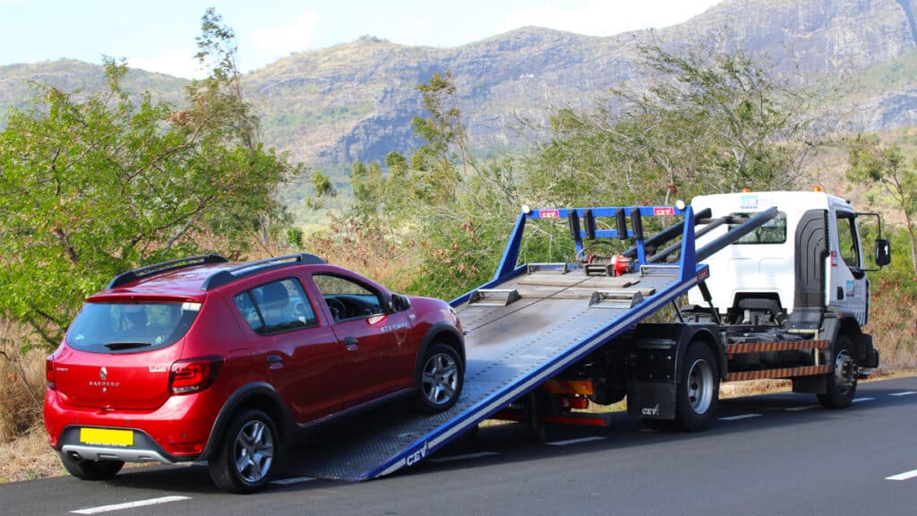 Red car being loaded onto a tow truck on a scenic mountainous road, with lush greenery in the background.