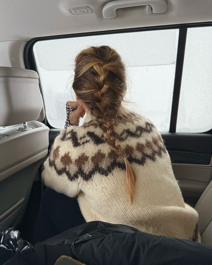 Woman with braid wearing a knitted sweater sits in a car, looking out a frosted window. Cozy winter travel scene.