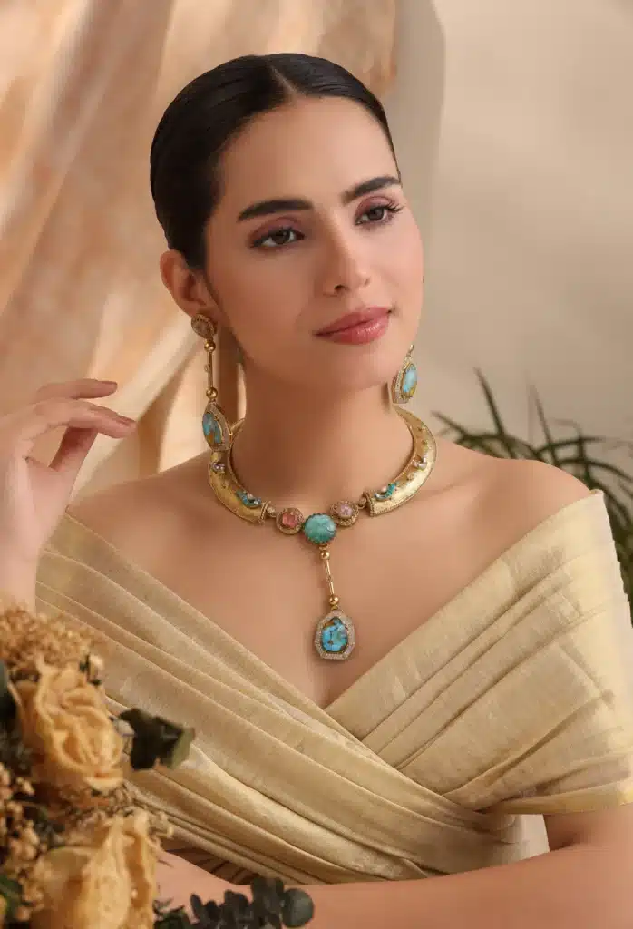 Elegant woman wearing gold and turquoise jewelry set with light beige off-shoulder dress, posing gracefully indoors.