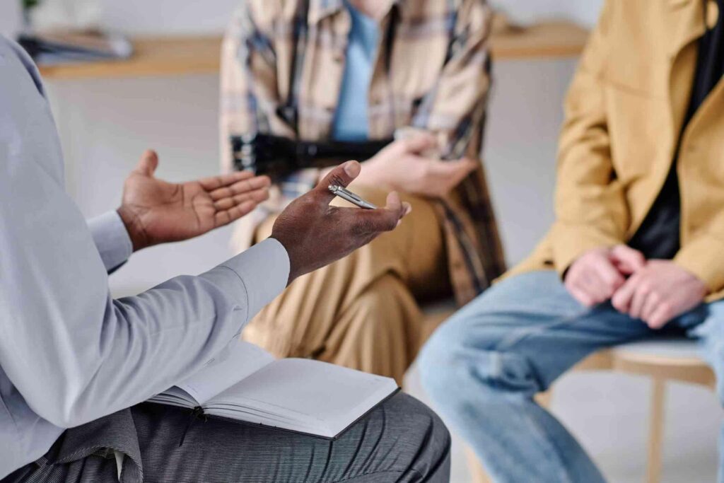 Therapist holds discussion with two individuals, open notebook, hands gesturing in conversation.