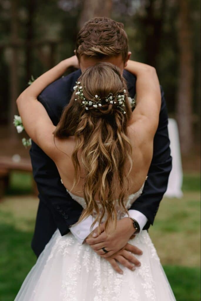 Bride and groom embrace, surrounded by nature, showcasing wedding dress and floral hairpiece in a heartfelt moment.