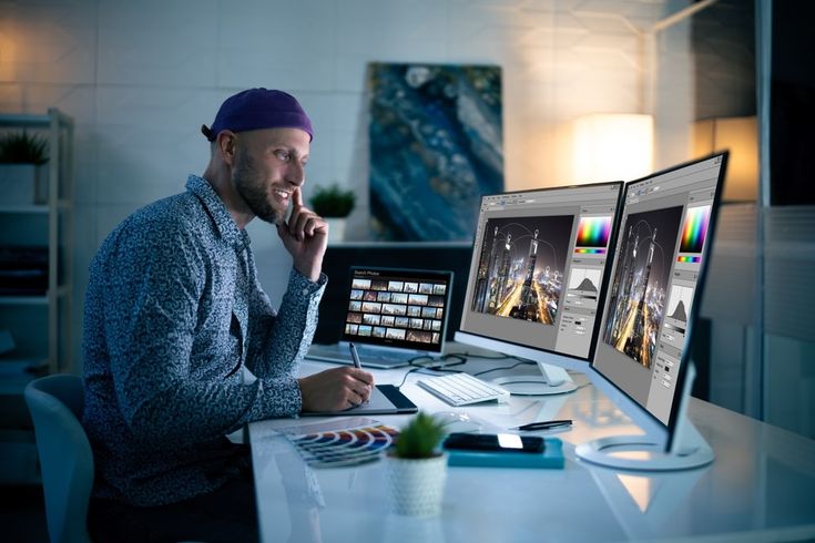 Graphic designer editing photos on dual monitors in a modern office, focused and creative workspace setup.