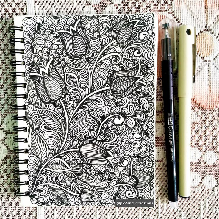 Intricate black and white floral doodle art on notebook with pens beside it. Perfect for creativity and inspiration.