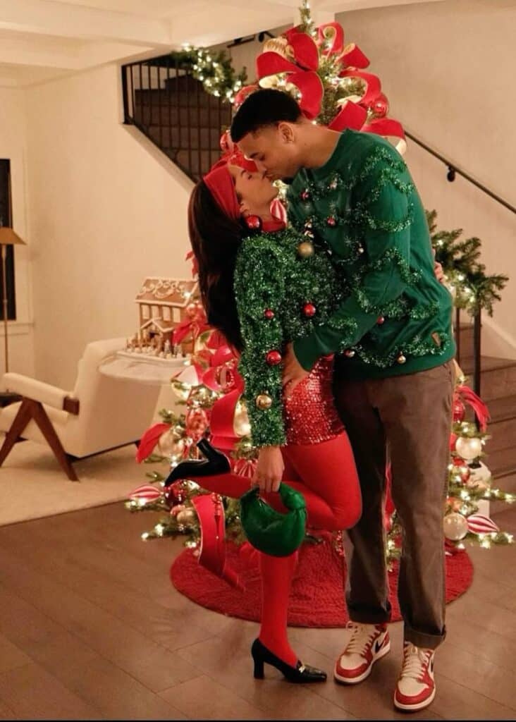 Couple in festive outfits kissing by decorated Christmas tree at home.