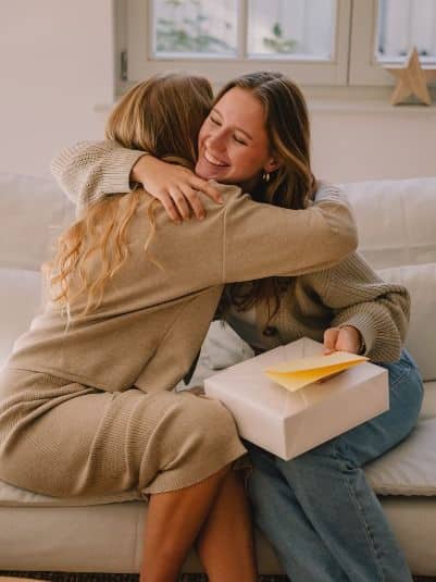 Two friends embrace on a couch, one holding a gift box with a card, expressing joy and friendship.