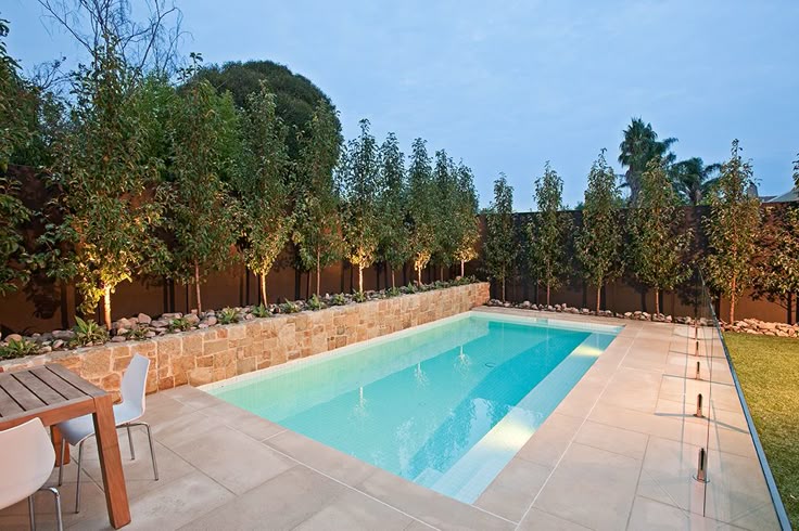 Modern backyard with illuminated swimming pool, surrounded by trees and patio furniture.