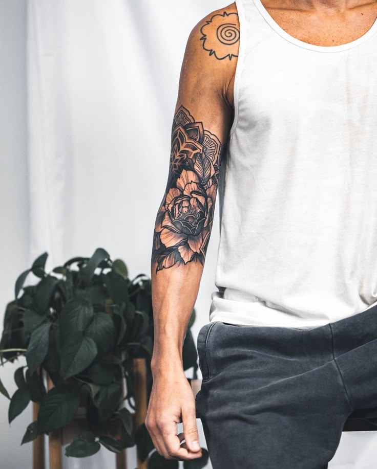 Man with floral and geometric arm tattoos wearing a white tank top, standing next to a potted plant.