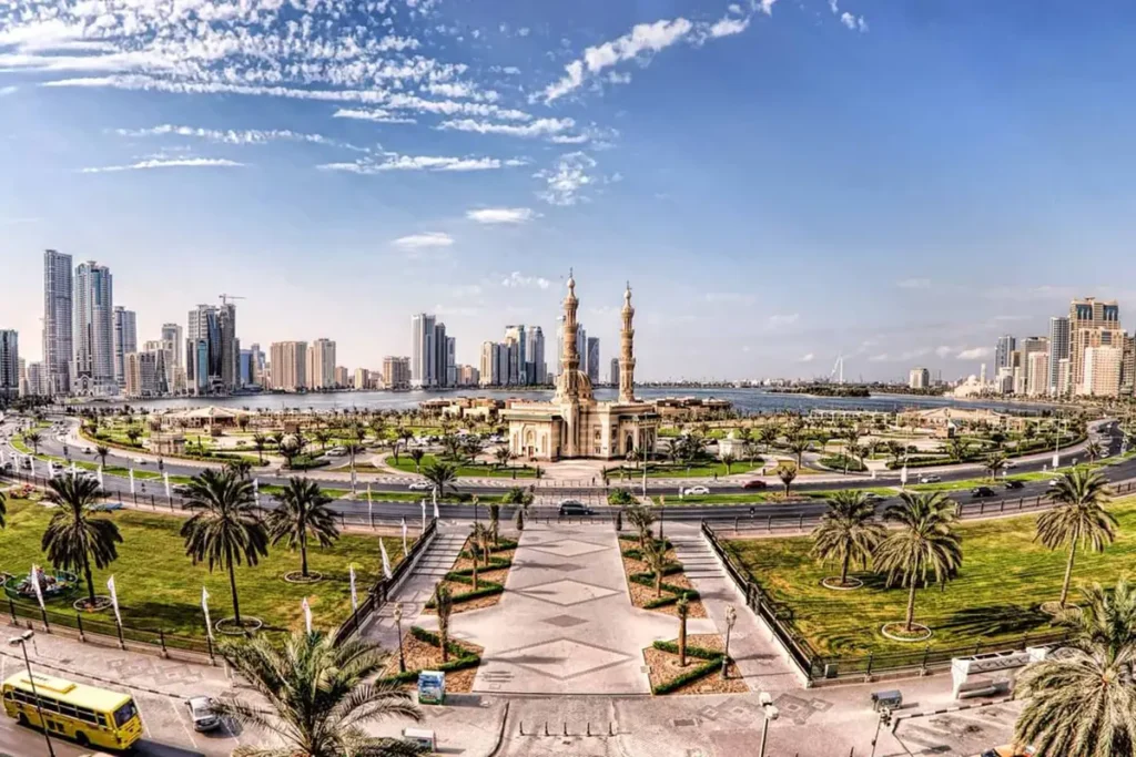 Panoramic view of Sharjah cityscape, featuring lush gardens, a central mosque, and tall modern skyscrapers under a clear sky.