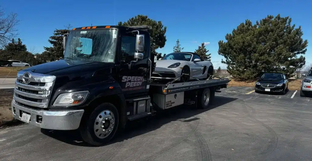 Tow truck carrying a sports car on a sunny day, parked beside trees and cars in a lot.