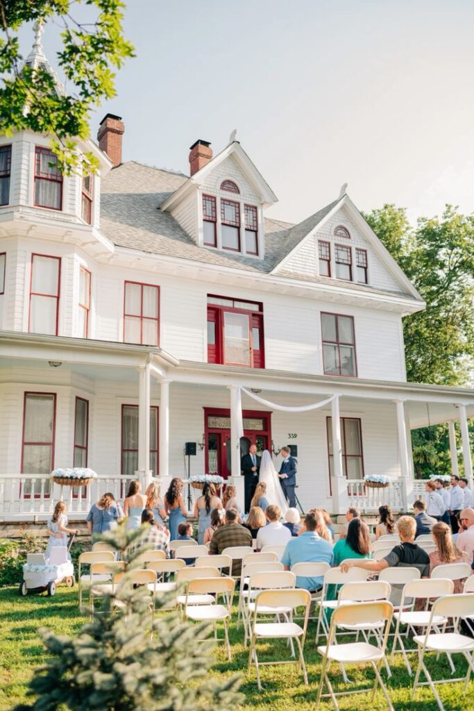 Outdoor wedding ceremony in front of a large white historic house with seated guests, a couple, and officiant.