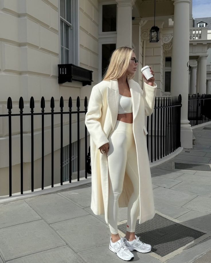 Woman in stylish white outfit holding coffee, standing in urban setting. Fashionable streetwear look with coat and sneakers.
