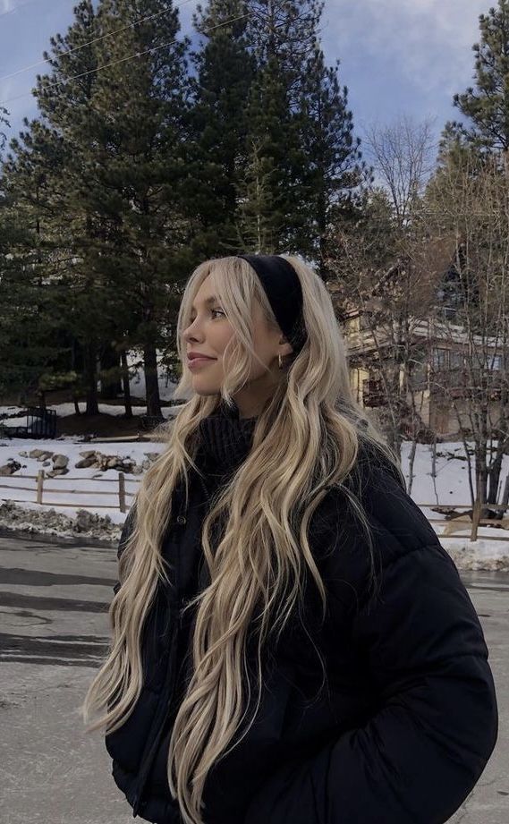 Woman with long blonde hair in winter jacket stands outdoors with snowy trees in background.
