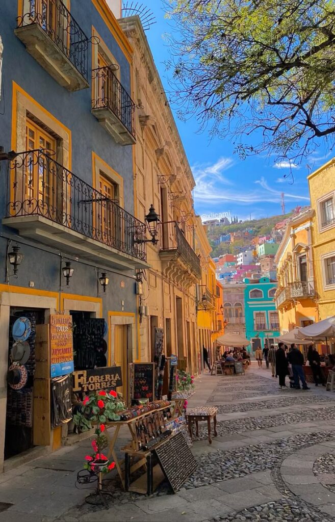 xiicami Colorful street in Guanajuato, Mexico, with vibrant buildings, cobblestones, and market stalls under a blue sky. | Sky Rye Design Colorful street in Guanajuato, Mexico, with vibrant buildings, cobblestones, and market stalls under a blue sky.