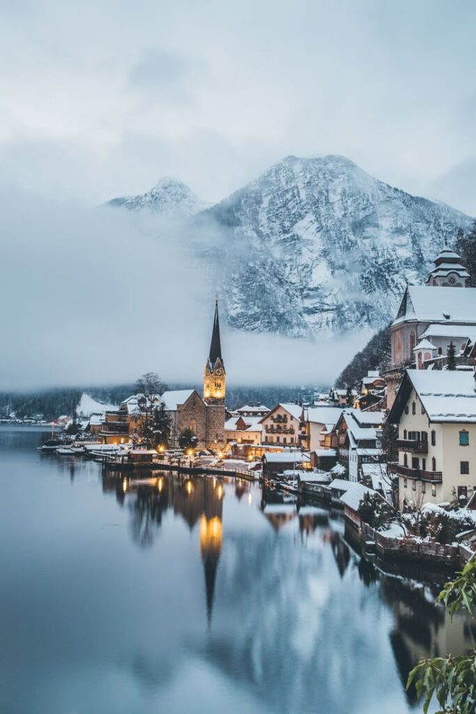 Австрия Snowy mountain village by a serene lake with church spire, mist, and cozy lights illuminating the snowy landscape. | Sky Rye Design Snowy mountain village by a serene lake with church spire, mist, and cozy lights illuminating the snowy landscape.