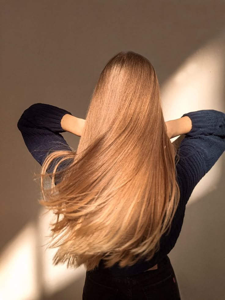 Woman with long, shiny blonde hair wearing a dark sweater, standing in sunlight, showcasing healthy and smooth hair.