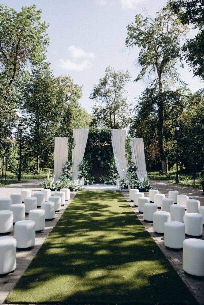 Outdoor wedding ceremony setup with white chairs and elegant floral archway on a green lawn aisle.