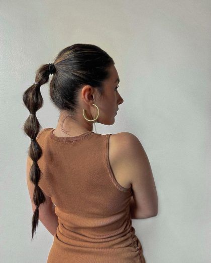 Woman with a bubble ponytail and hoop earrings in a brown tank top, facing away from the camera against a neutral background.