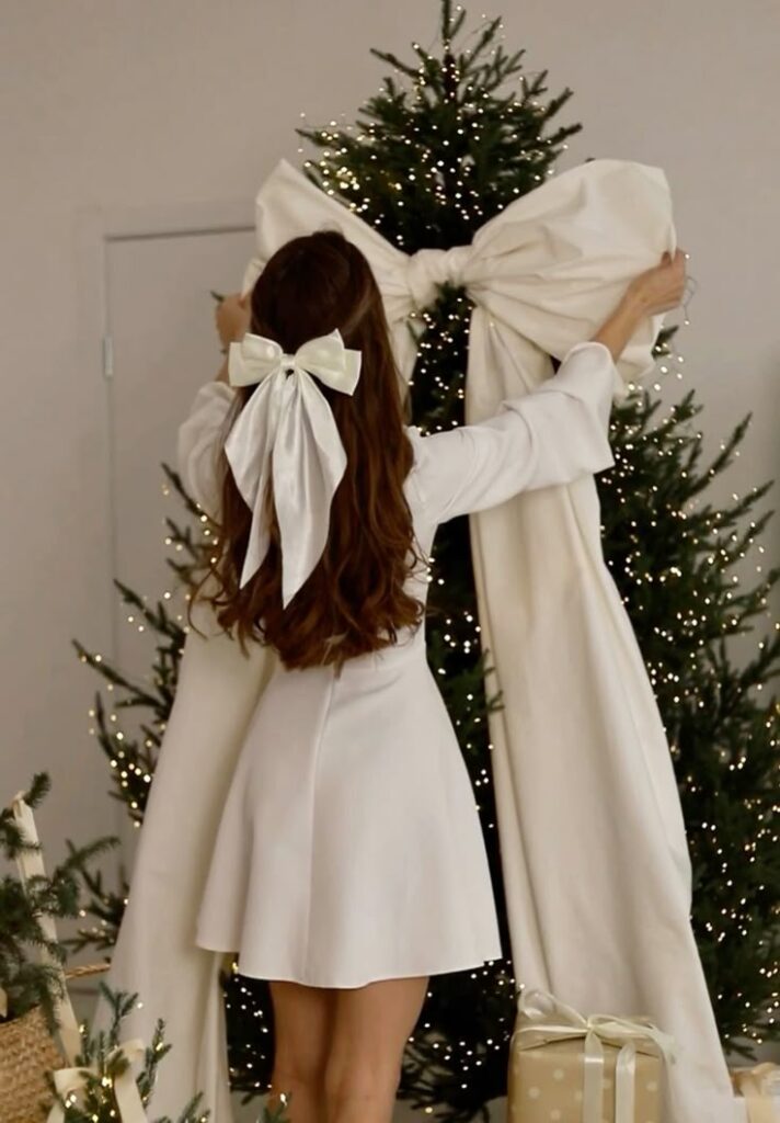 Woman in white dress decorating Christmas tree with large ribbon bow, festive gifts underneath.