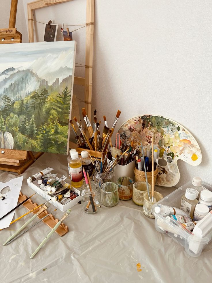 Art studio with landscape painting, brushes, paint tubes, and palette on a table covered with a protective cloth.
