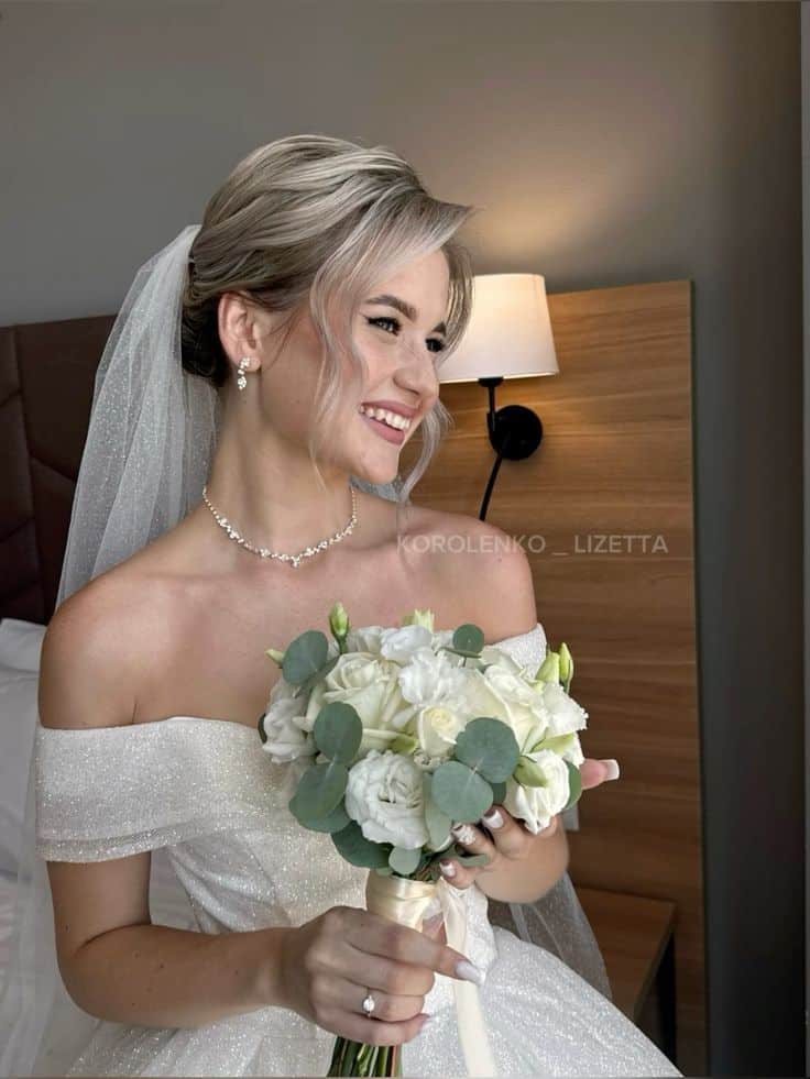 Bride in off-shoulder dress smiles, holding bouquet of white roses and greenery, standing indoors.