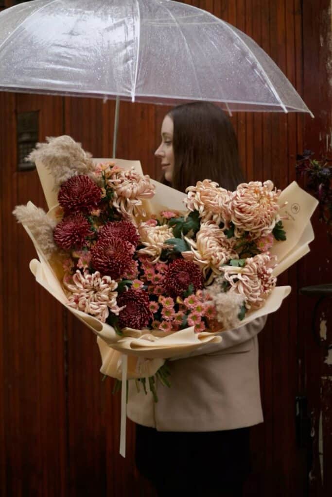 Woman holding a large bouquet of flowers under a transparent umbrella, standing against a wooden door background.