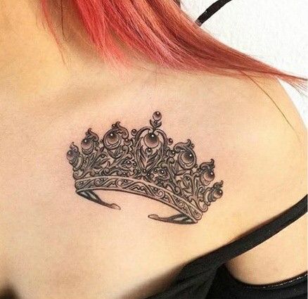 завантаження Intricate crown tattoo on a person's shoulder, under red hair, showcasing detailed black ink design. | Sky Rye Design Intricate crown tattoo on a person's shoulder, under red hair, showcasing detailed black ink design.