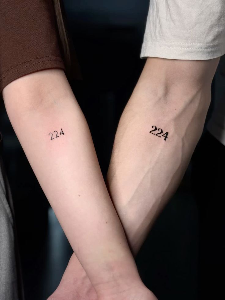 Matching 224 tattoos on crossed forearms, symbolizing a shared connection or significant date.