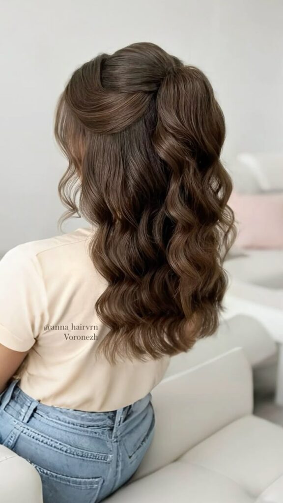 Elegant wavy brunette hairstyle, half-updo on a woman wearing a cream top and jeans, showcasing soft curls.