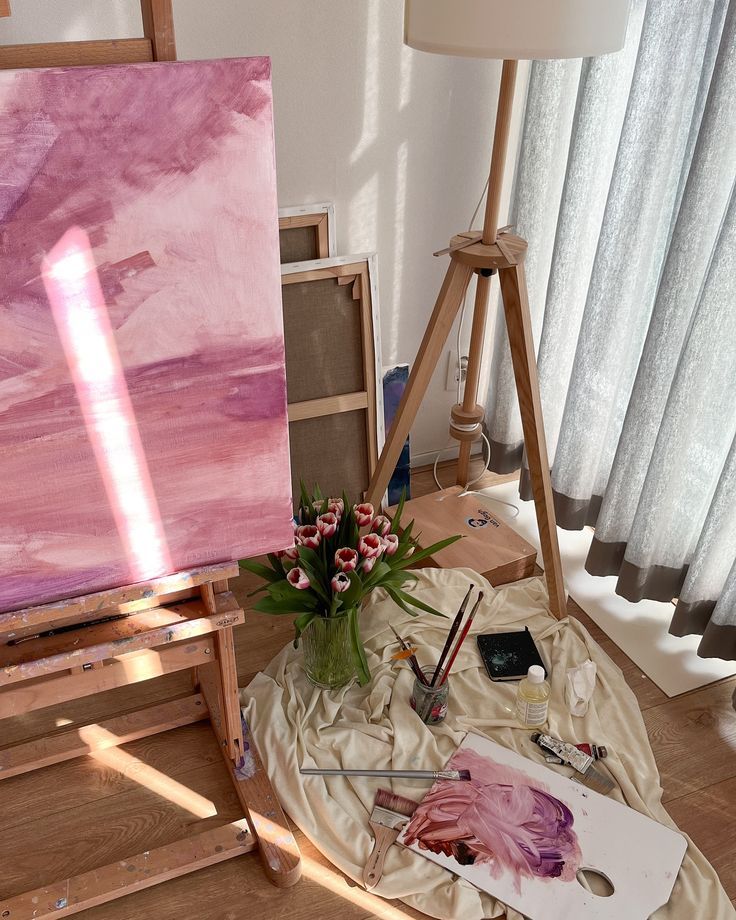 Art studio setup with pink painting, tulips, and painting supplies on a wooden floor by a window.