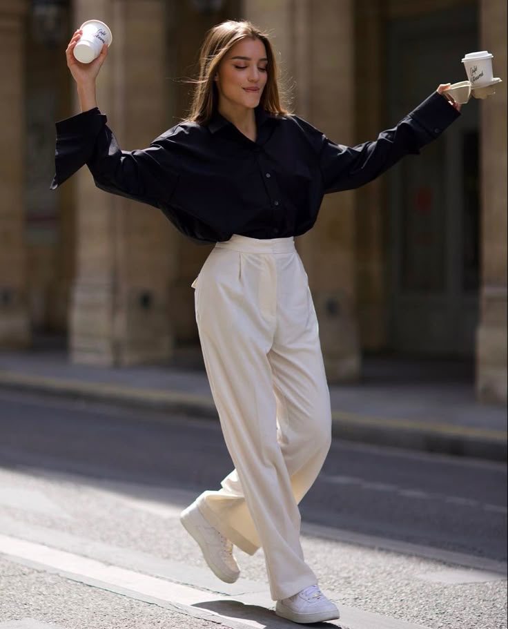 ☕️ Woman joyfully balancing coffee cups while walking on a city street, wearing a black blouse and white pants. | Sky Rye Design Woman joyfully balancing coffee cups while walking on a city street, wearing a black blouse and white pants.