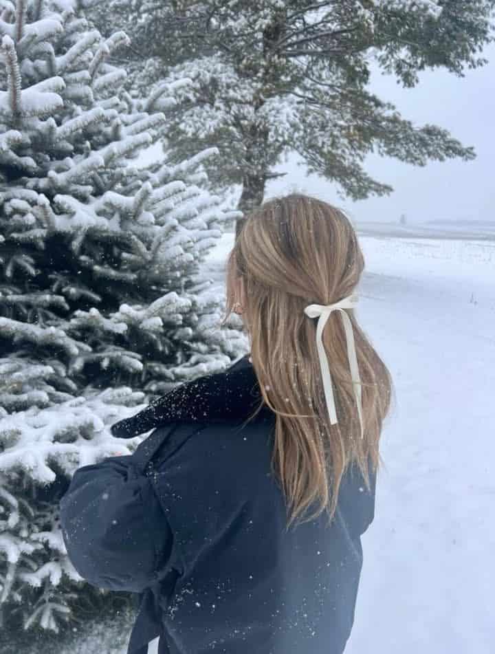 Person in winter coat and ribbon hair bow by snow-covered pine tree in snowy landscape.