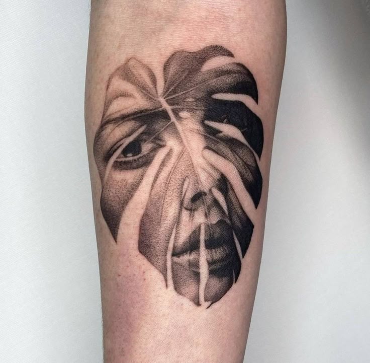 Tattoo of a face intertwined with a monstera leaf on forearm, showcasing artistic blending and intricate design.