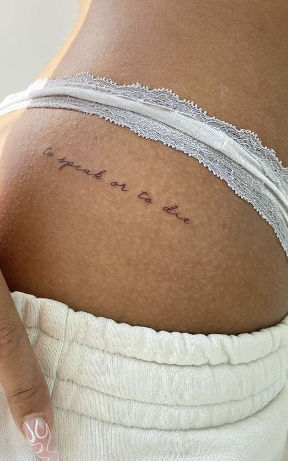 Small script tattoo on hip saying to speak or to die, above white lace underwear, showing personal expression.