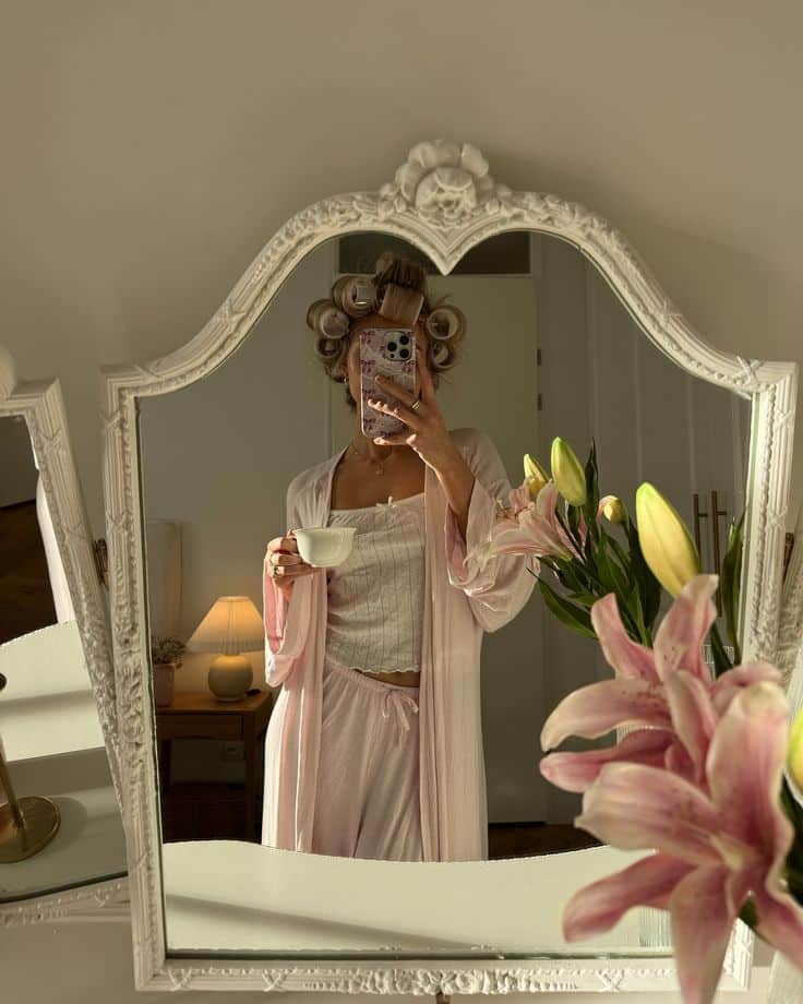 Woman in pink robe with hair curlers, drinking coffee in front of ornate mirror, with lilies in foreground.