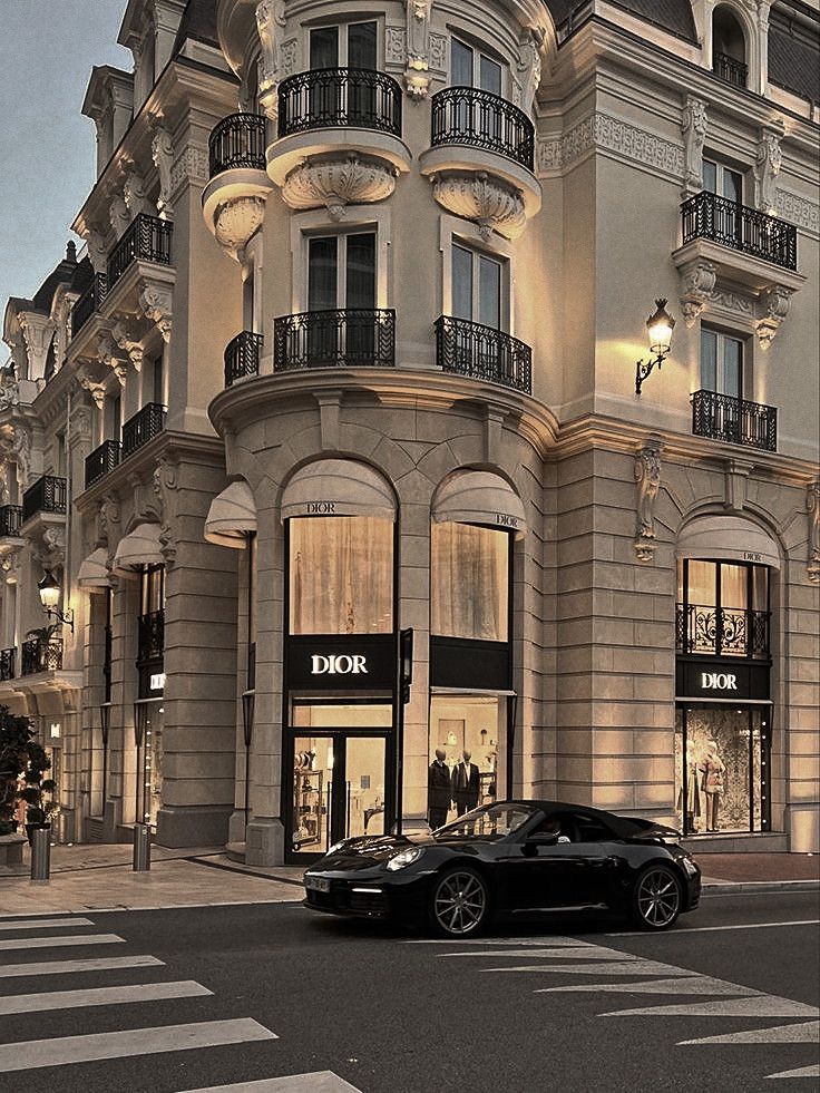 ✦┈๑⋅⋯ ✨ ⋯⋅๑┈✦ 💸☕🖤 ✧ Please do not repeat Elegant Dior storefront on a city corner with a sleek black car parked at dusk. | Sky Rye Design Elegant Dior storefront on a city corner with a sleek black car parked at dusk.