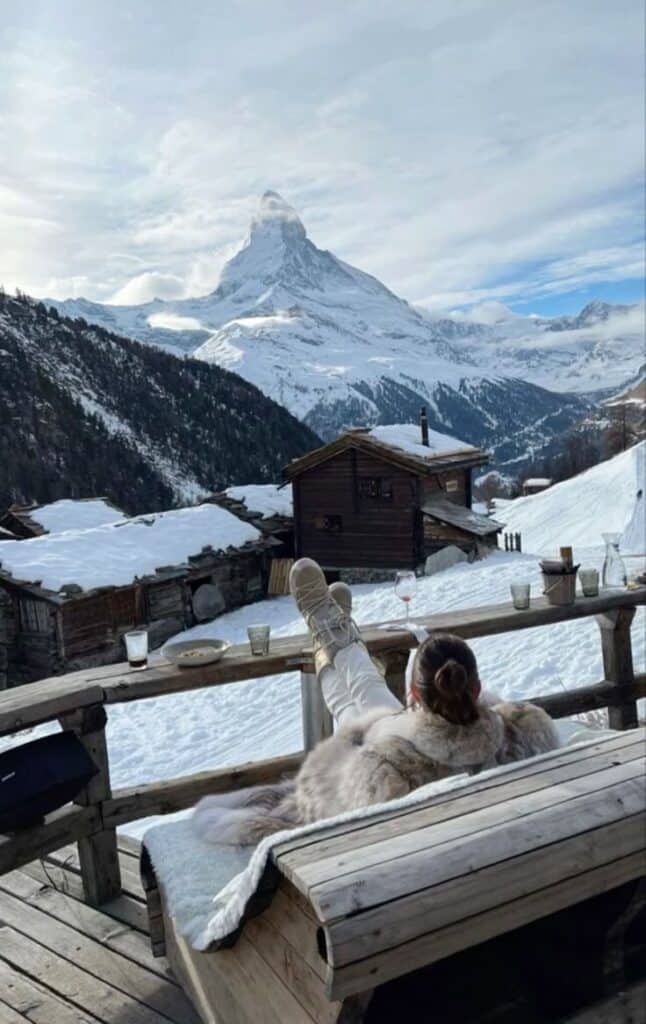❄️ Person relaxing on a snowy mountain terrace, enjoying a scenic view of the Matterhorn with rustic cabins in the foreground. | Sky Rye Design Person relaxing on a snowy mountain terrace, enjoying a scenic view of the Matterhorn with rustic cabins in the foreground.