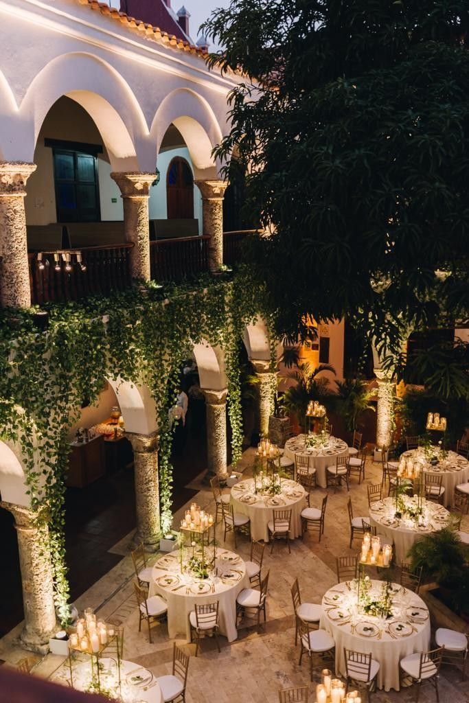 Elegant courtyard wedding setup with round tables, floral centerpieces, gold chairs, candles, and archways draped in leafy greenery.