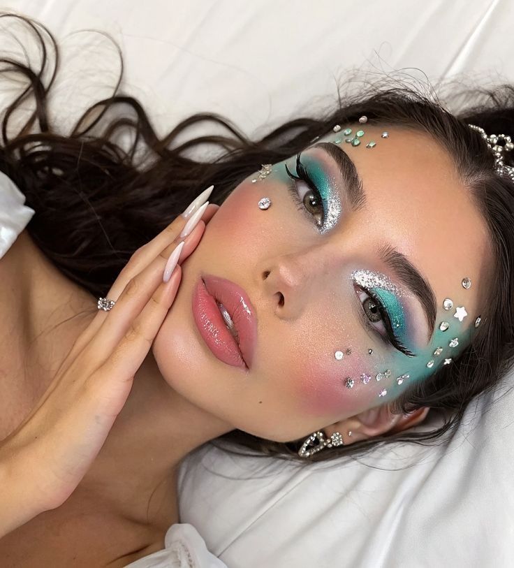 Woman with turquoise and silver eye makeup, gem accents, and glossy lips, posing on a white background. Fantasy-inspired beauty look.