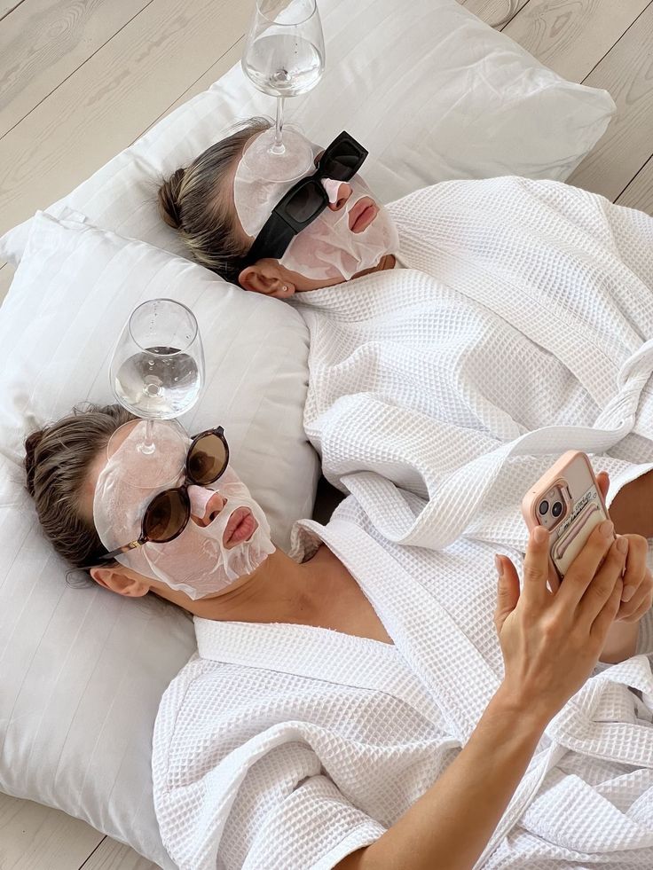 Women relaxing with sheet masks, wearing sunglasses and robes, balancing wine glasses on their heads, taking a selfie.