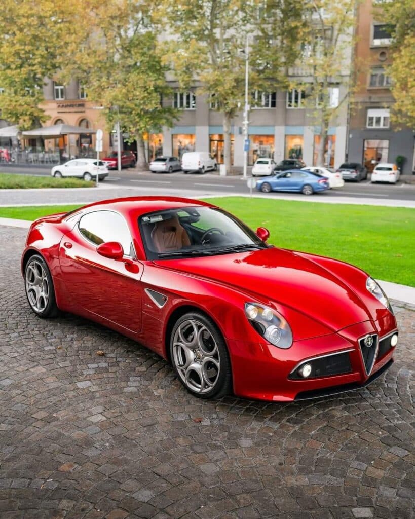 02031d436a8a97838dfb82e0682fcde9 Sleek red sports car parked on a cobblestone street, surrounded by greenery and urban buildings in the background. | Sky Rye Design Sleek red sports car parked on a cobblestone street, surrounded by greenery and urban buildings in the background.