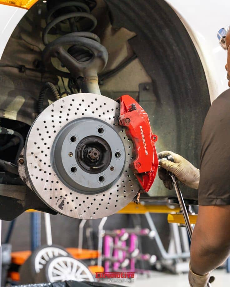 022698de5a59d67a37114f149cfd7c43 Mechanic working on high-performance car brake system, featuring a red brake caliper and drilled rotor under a lifted vehicle. | Sky Rye Design Mechanic working on high-performance car brake system, featuring a red brake caliper and drilled rotor under a lifted vehicle.