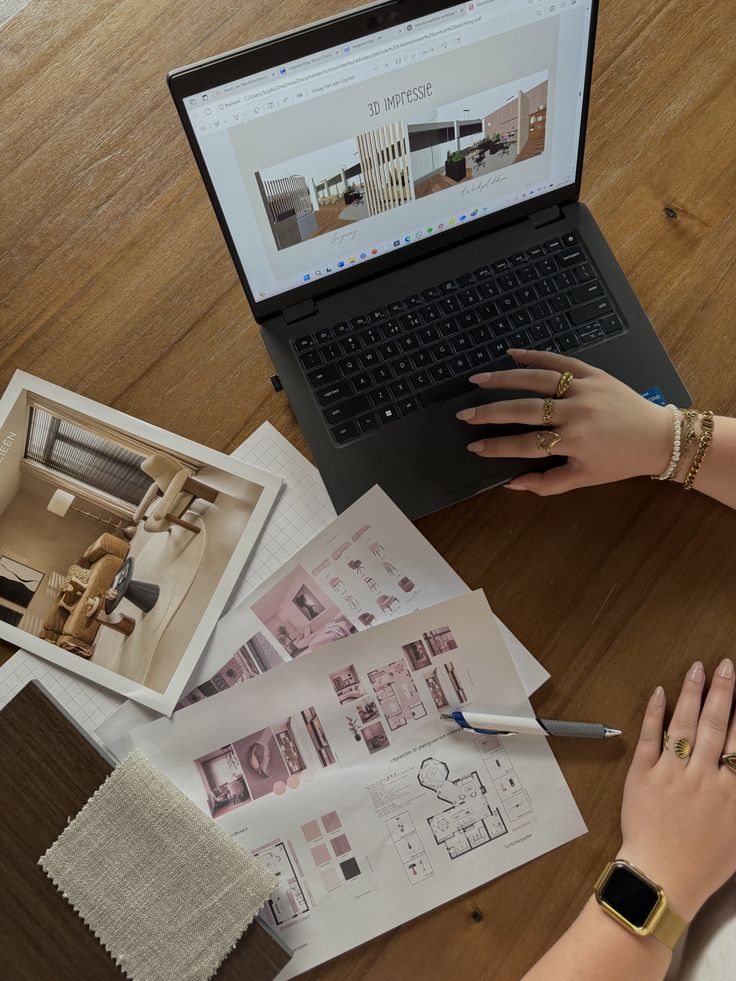 Person working on interior design projects with laptop, floor plans, and material samples on a wooden table.