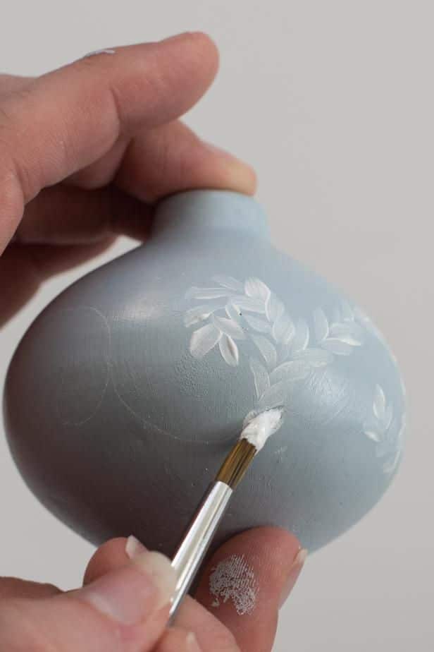 Hand painting white floral design on small blue vase with brush.