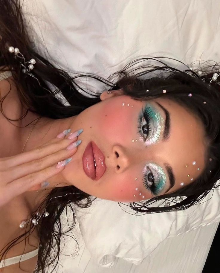 Woman with vibrant turquoise eyeshadow and rhinestone face accents lying on a pillow, showcasing glam makeup and long painted nails.