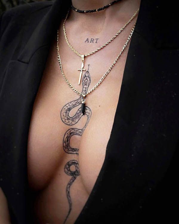 Close-up of a snake tattoo on a person’s chest with layered necklaces and a black blazer, featuring the word ART above the design.